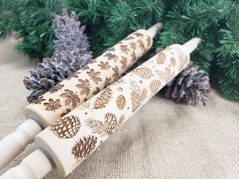 May include: Two wooden rolling pins with engraved designs. One pin features a repeating pattern of maple leaves, while the other has a pattern of pine cones and branches.