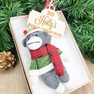 May include: A gray sock monkey ornament wearing a green sweater and red scarf. The ornament is in a white box with a wooden tag that reads "2019 Holly's First Christmas".