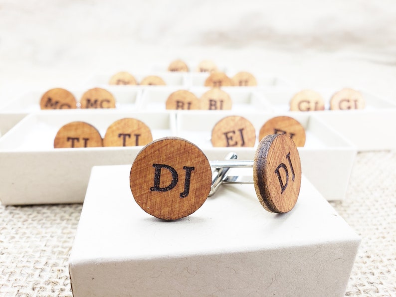 May include: A pair of wooden cufflinks with the initials "DJ" engraved on them. The cufflinks are in a white box and are surrounded by other cufflinks with different initials.