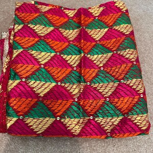 May include: A folded textile with a vibrant geometric pattern. The design features diamond shapes in shades of pink, orange, green, and gold, with small embellishments. The fabric appears to be intricately woven, showcasing a traditional craft.