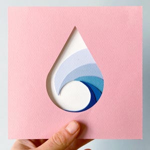 May include: A pink square paper cutout featuring a teardrop-shaped void, showcasing a layered paper design of a wave in varying shades of blue. The wave design is constructed from curved paper strips, producing a 3D effect. The artwork is held by a hand.