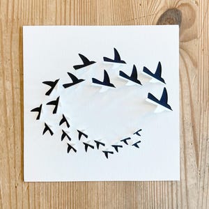 May include: A white square card features a flock of black and white birds in flight, arranged in a circular pattern. The birds are cut out, creating a layered effect. The background is a light wood grain.