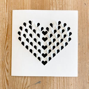 May include: A white card with a black and white heart made of paper hearts. The heart is made of many small hearts, each half black and half white.