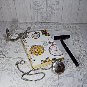 Könnte beinhalten: Ein weißes Notizbuch mit einem Uhr- und Zahnrad-Design. Das Notizbuch ist geöffnet und hat eine silberne Kette daran befestigt. Auf einer weißen Spitzenunterlage liegen zwei silberne Taschenuhren mit Ketten. Ein schwarzer Stift liegt auf der Unterlage.