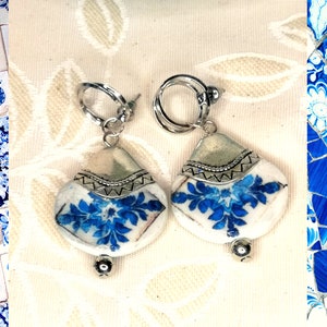 May include: A pair of silver hoop earrings with blue and white ceramic teardrop-shaped pendants. The pendants feature a floral design.