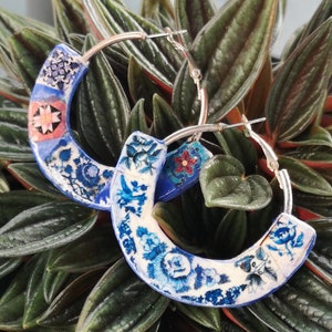Op de afbeelding: Een paar blauwe en witte creolen met een bloemenpatroon. De oorbellen zijn gemaakt van keramiek en hebben een zilveren metalen ring.