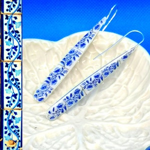 May include: A pair of long, white earrings with a blue and white floral pattern. The earrings are made of ceramic and have a silver wire.