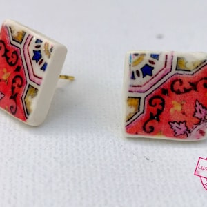 May include: A pair of square ceramic earrings with a colorful geometric pattern. The earrings are white with a red, pink, blue, and yellow pattern.