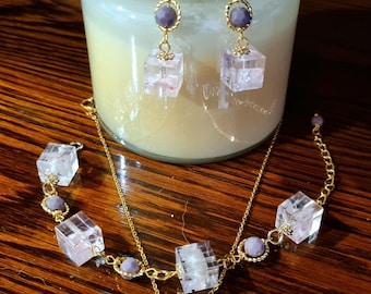 Puede incluir: Un conjunto de joyas de tono dorado con un collar y pendientes. El collar y los pendientes tienen colgantes transparentes en forma de cubo y pequeñas cuentas redondas de color púrpura. Los pendientes tienen un gancho de tono dorado. Las joyas se exhiben sobre una superficie de madera.