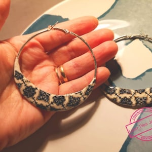 Può includere: Un paio di grandi orecchini a cerchio argentati con un motivo geometrico bianco e nero. Gli orecchini sono in metallo e hanno una chiusura a cerniera.