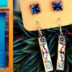 May include: A pair of gold dangle earrings with blue and white ceramic tiles featuring floral patterns. The tiles are rectangular and have a gold trim. The earrings are displayed on a brown card.
