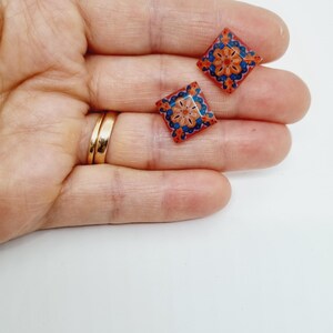 K&ouml;nnte beinhalten: Ein Paar kleine, quadratische Ohrringe mit einem bunten geometrischen Muster. Die Ohrringe sind rot, orange, blau und wei&szlig;.