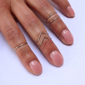 May include: A set of four silver rings, two simple bands and two chevron shaped rings, worn on a hand with nude nail polish.