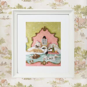 May include: A framed print depicts a cozy scene of a person reading in bed, surrounded by several dogs. The bed has a pink headboard and a green patterned background. A tray with food and drinks sits on the bed. The frame is white.