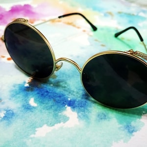 Puede incluir: Gafas de sol redondas con montura dorada y lentes negras sobre un fondo de acuarela.