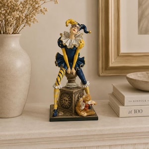 May include: A decorative figurine of a jester in a blue and yellow outfit, perched atop a pedestal. The jester has a white ruffled collar and a whimsical hat. A small teddy bear sits at the base of the pedestal. The figurine is a decorative item.