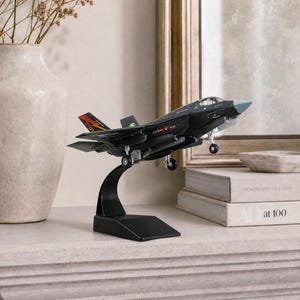May include: A black model airplane with a gray nose and red and yellow accents is displayed on a black stand. The model is a detailed replica of a fighter jet, showcasing its wings, tail, and landing gear. The model is set against a neutral background.