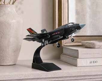 May include: A black model airplane with a gray nose and red and yellow accents is displayed on a black stand. The model is a detailed replica of a fighter jet, showcasing its wings, tail, and landing gear. The model is set against a neutral background.