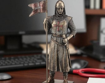 May include: A bronze-colored statue of a medieval knight holding a spear with a small flag. The knight is wearing a helmet, chainmail, and a tunic with a red cross. The statue is standing on a rectangular base.