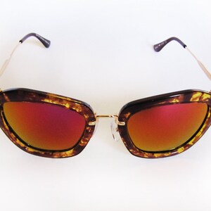 May include: Cat-eye sunglasses with a tortoiseshell frame and gold-tone metal arms. The lenses have a gradient red and orange tint. The sunglasses are set against a white background.