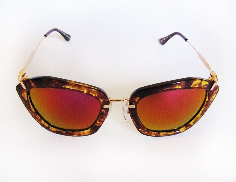 May include: Cat-eye sunglasses with a tortoiseshell frame and gold-tone metal arms. The lenses have a gradient red and orange tint. The sunglasses are set against a white background.