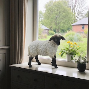 May include: A ceramic sheep figurine with a black face, ears, and legs, and a white, textured body. The sheep is standing on a white surface in front of a window, with a view of a green yard and trees.