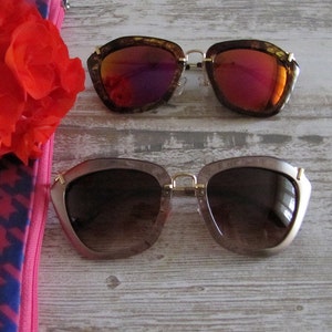 May include: Two pairs of sunglasses with gold frames and unique, angular shapes. One pair has brown frames with red-tinted lenses, and the other has light brown frames with brown-tinted lenses. A red flower and a pink and blue patterned pouch are also visible.