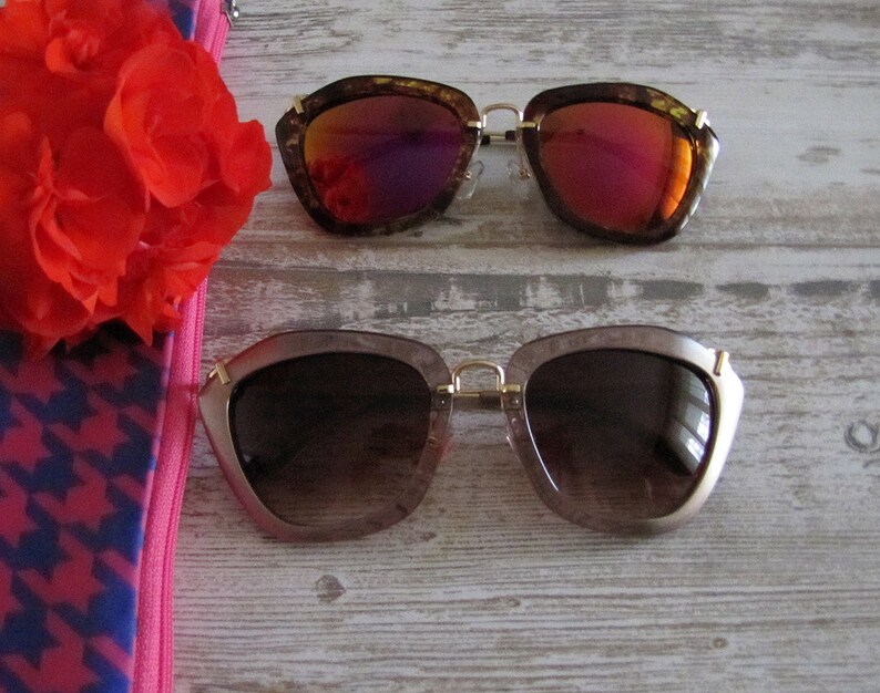 May include: Two pairs of sunglasses with gold frames and unique, angular shapes. One pair has brown frames with red-tinted lenses, and the other has light brown frames with brown-tinted lenses. A red flower and a pink and blue patterned pouch are also visible.