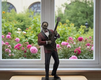 May include: A statue of a musician playing a guitar. The figure is wearing a burgundy blazer, black pants, and a bow tie. The guitar is black. The statue is on a dark base, set against a window with a garden view.