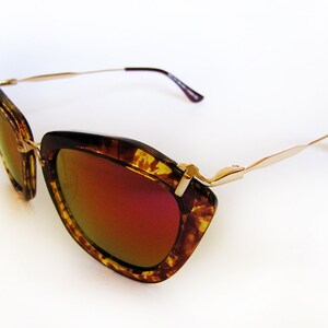May include: Cat-eye sunglasses with a tortoiseshell pattern frame and gold-tone metal arms. The lenses have a mirrored, reddish-orange tint. The sunglasses are set against a white background.