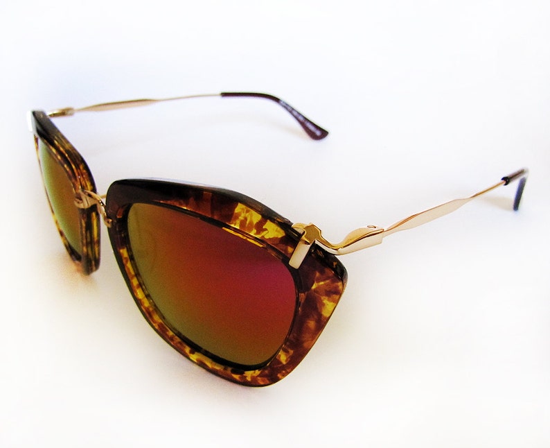 May include: Cat-eye sunglasses with a tortoiseshell pattern frame and gold-tone metal arms. The lenses have a mirrored, reddish-orange tint. The sunglasses are set against a white background.