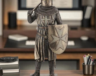 May include: A detailed figurine of a medieval knight in full armor, holding a shield with a cross design and a weapon. The knight's helmet has a cross-shaped visor. The figurine is bronze-colored and stands on a rectangular base.