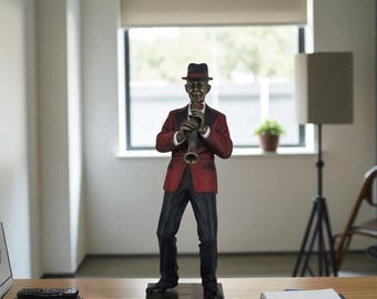 May include: A bronze-colored statue of a jazz musician playing a clarinet. The figure wears a red blazer, black pants, and a fedora. The statue is on a square base with a checkered pattern. The background includes a window, a lamp, and a desk.