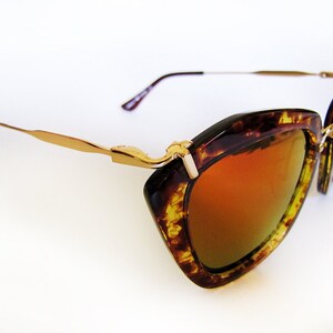 May include: Cat-eye sunglasses with a tortoiseshell frame and gold-tone metal accents. The lenses have a gradient orange tint. The temples are thin and gold-colored, with a dark brown earpiece. The sunglasses are set against a white background.