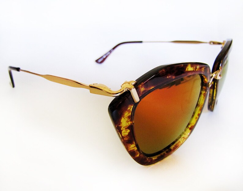 May include: Cat-eye sunglasses with a tortoiseshell frame and gold-tone metal accents. The lenses have a gradient orange tint. The temples are thin and gold-colored, with a dark brown earpiece. The sunglasses are set against a white background.