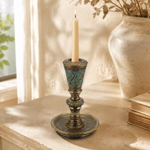 May include: A metallic candlestick holder with a wide circular base, a tapered stem, and a flared top cup. The surface features an intricate pattern of blue and gold filigree detailing. A single cream-colored taper candle is lit inside the holder.
