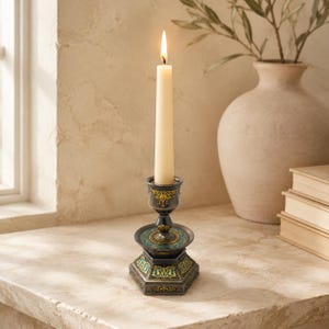 May include: A lit cream-colored taper candle sits in a dark metal candlestick holder. The holder features an octagonal base and a circular drip tray, both decorated with intricate teal and gold filigree patterns against a dark metallic finish.