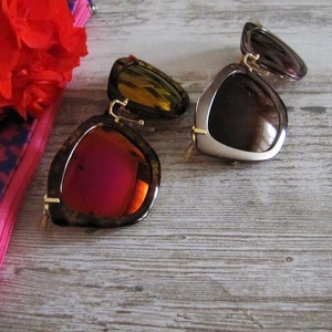 May include: Two pairs of sunglasses with unique frames and lenses. One pair features a tortoiseshell frame with red lenses, while the other has a brown frame with brown lenses. Both have gold-colored accents. A red flower and a pink and blue patterned case are in the background.