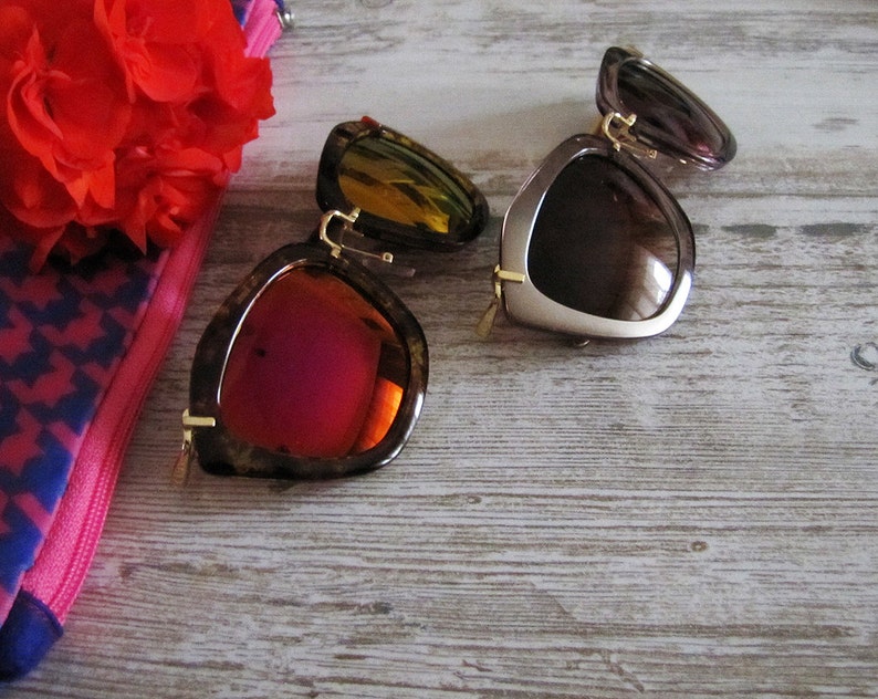 May include: Two pairs of sunglasses with unique frames and lenses. One pair features a tortoiseshell frame with red lenses, while the other has a brown frame with brown lenses. Both have gold-colored accents. A red flower and a pink and blue patterned case are in the background.