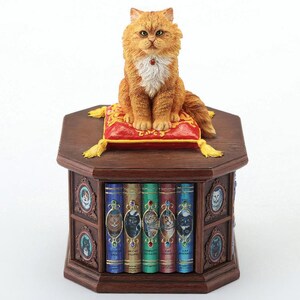 May include: A detailed figurine of a ginger cat with a white chest, sitting atop a red cushion with gold tassels. The cat is perched on a dark wooden cabinet with decorative books featuring cat portraits.