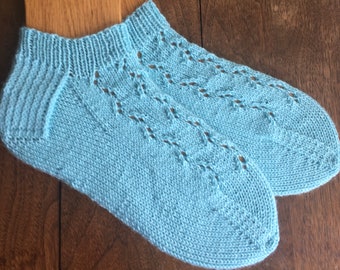 May include: A pair of light blue knitted ankle socks. The socks have a ribbed cuff and a decorative openwork pattern along the top. The socks are displayed on a wooden surface.