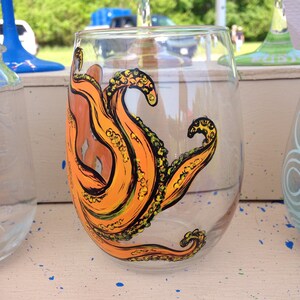 May include: A clear stemless wine glass with an orange octopus tentacle design. The tentacles are painted with black outlines and yellow details. The glass is part of a set, with other glasses visible in the background.