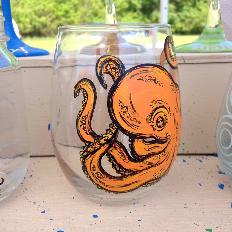 May include: A stemless wine glass with an orange octopus design. The octopus is painted with black outlines and yellow accents. The glass is clear, and the background is blurred.