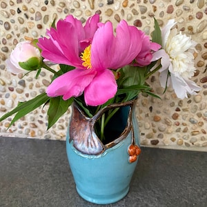 Op de afbeelding: Een blauwe keramische vaas met een bruin bladontwerp en een klein cluster rode bessen. De vaas is gevuld met roze en witte pioenen.