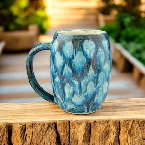 May include: A ceramic mug with a blue and teal pattern. The mug has a large handle and a wide opening. The design features a repeating pattern of teardrop shapes in varying shades of blue. The mug is sitting on a wooden surface.