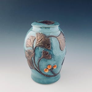 May include: A teal ceramic urn with a lid, decorated with bronze-coloured ginkgo leaf designs and small orange berries. The urn has a glossy finish and a rounded shape, suitable for memorial purposes.