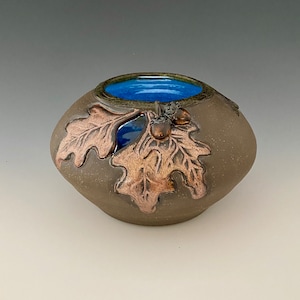 May include: A small, brown ceramic pot with a blue glaze inside. The pot is decorated with a raised design of oak leaves and acorns.