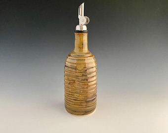 Puede incluir: Una botella de cerámica para aceite de oliva con un pico vertedor de acero inoxidable. La botella tiene un diseño a rayas horizontales texturizado en tonos marrones y beige. El pico vertedor es plateado y tiene una tapa con bisagras.
