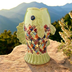 May include: A green ceramic fish-shaped vase with a colourful scale pattern. The vase has fins and a large eye. It is set on a rock, with a mountain range in the background. The vase is approximately 25 cm tall.