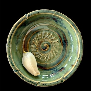 Może przedstawiać: Ręcznie wykonana, zielona ceramiczna tarka do czosnku ze spiralnym wzorem pośrodku. Tarka ma teksturowaną krawędź, a na powierzchni spoczywa jasny ząbek czosnku. Tło jest czarne.